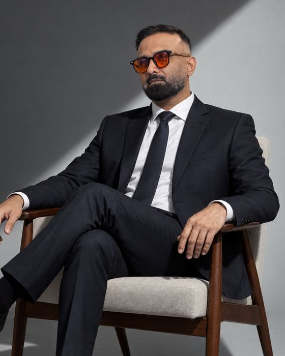 Dr Krish Bharggav in a black suit and tie, seated in an armchair in a grey studio